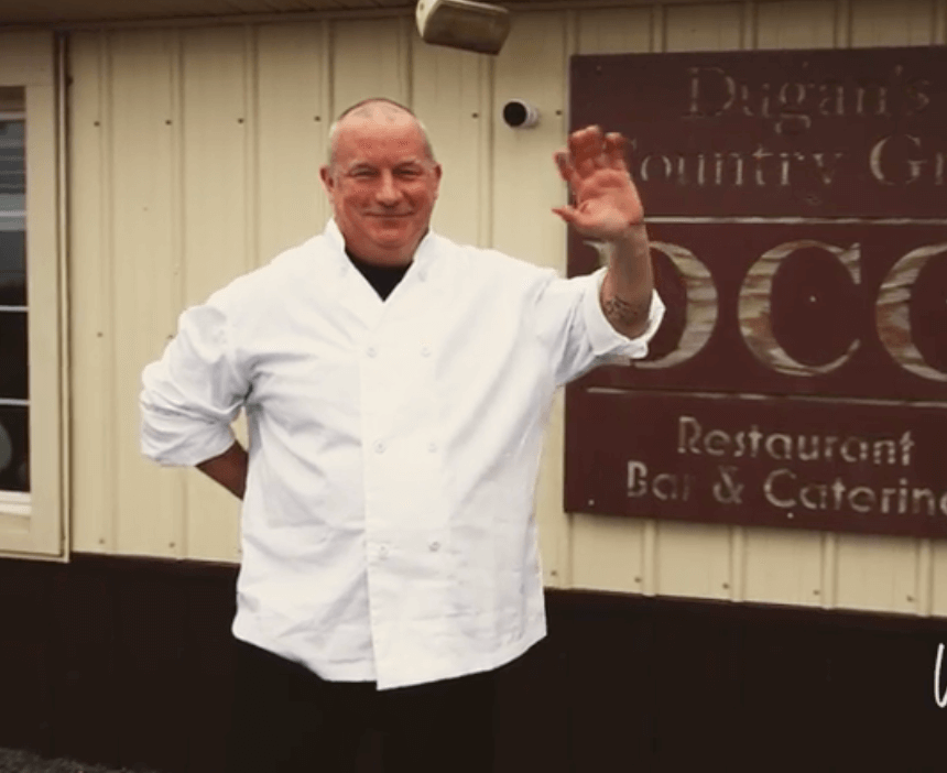 Photo of a male chef with a white chef coat on standing and waving outside of restaurant